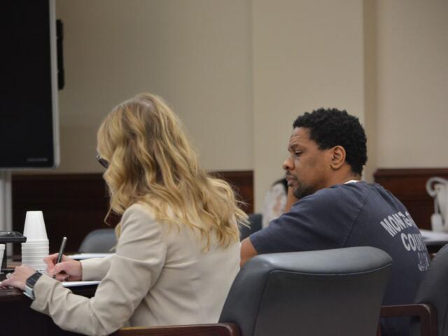 Two people sit at a conference table in a formal room; a blonde woman in a beige jacket writes in a notebook while a man in a dark T-shirt sits beside her.