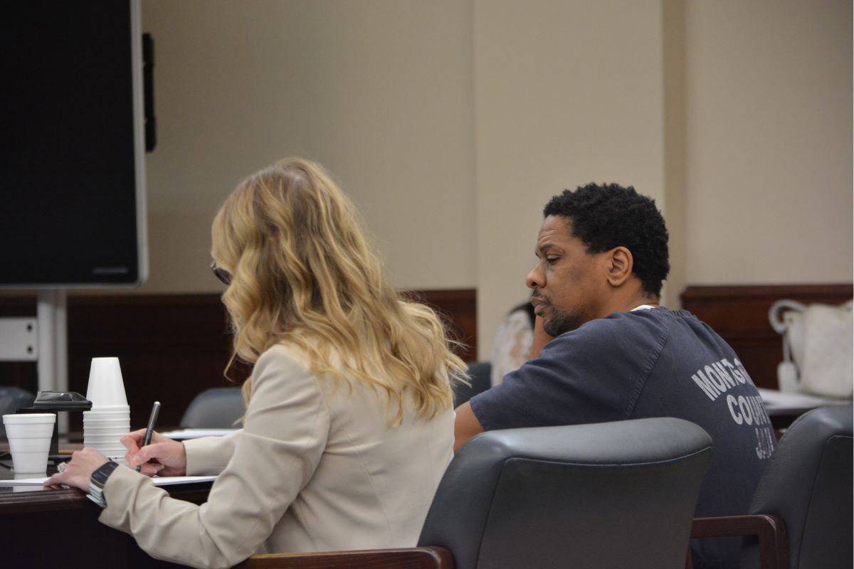 Two people sit at a conference table in a formal room; a blonde woman in a beige jacket writes in a notebook while a man in a dark T-shirt sits beside her.