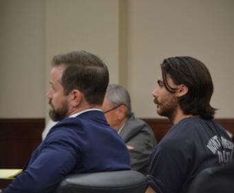 Two men sit side by side in a courtroom, one in a blue suit and the other with long hair in a dark shirt, facing forward.