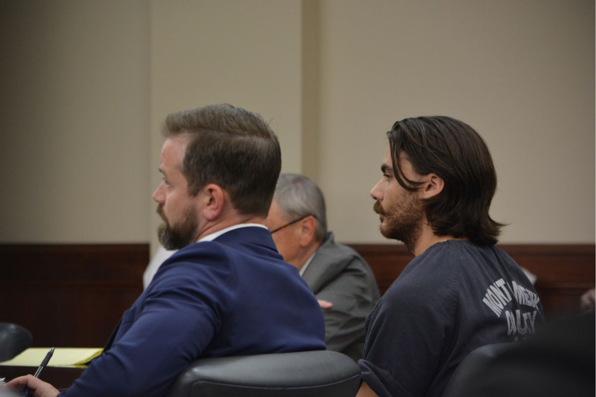 Two men sit side by side in a courtroom, one in a blue suit and the other with long hair in a dark shirt, facing forward.