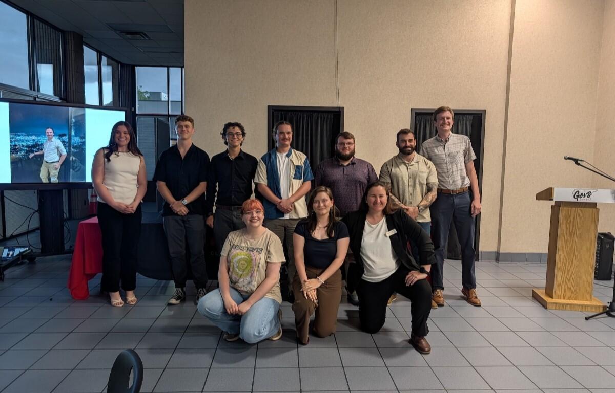 Group of ten people posing for a photo in a conference room with a presentation screen on the left and a wooden podium nearby.