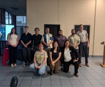 Group of ten people posing for a photo in a conference room with a presentation screen on the left and a wooden podium nearby.