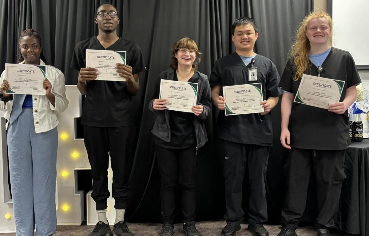 Five graduates stand on a stage holding certificates and smiling, against a black curtain backdrop.