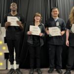 Five graduates stand on a stage holding certificates and smiling, against a black curtain backdrop.