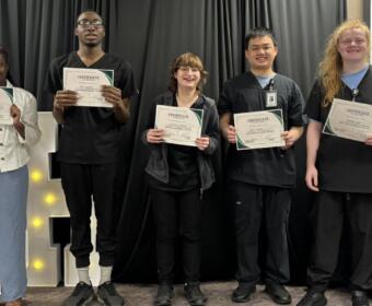 Five graduates stand on a stage holding certificates and smiling, against a black curtain backdrop.