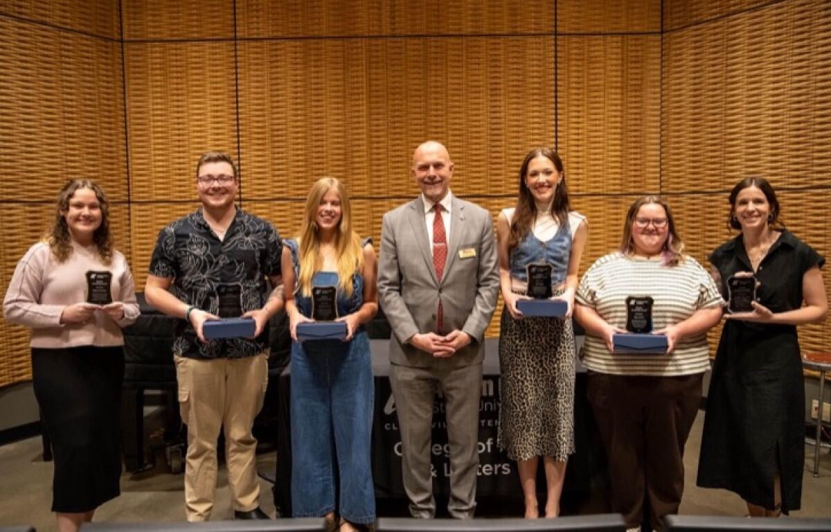 Seven award recipients stand in a row on a stage, each holding a glass plaque.