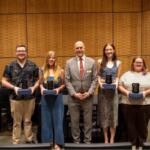 Seven award recipients stand in a row on a stage, each holding a glass plaque.
