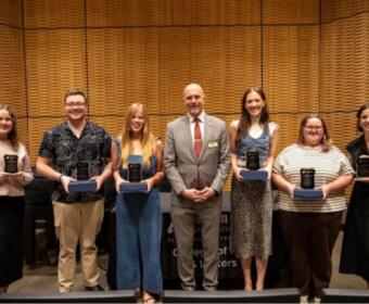 Seven award recipients stand in a row on a stage, each holding a glass plaque.