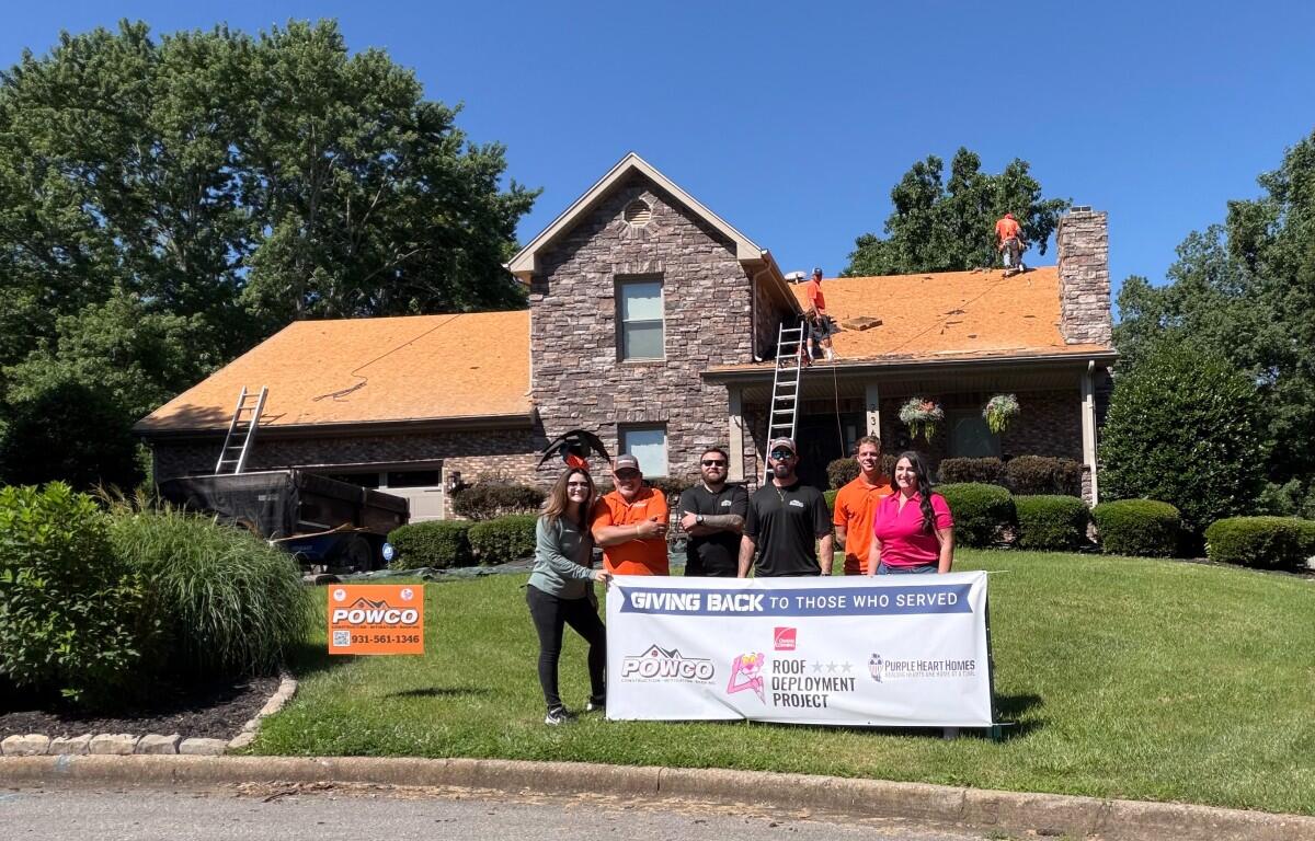 Volunteer group posing with a banner in front of a stone house while roof installers work on the orange roof on top of the house.