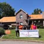 Volunteer group posing with a banner in front of a stone house while roof installers work on the orange roof on top of the house.