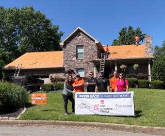 Volunteer group posing with a banner in front of a stone house while roof installers work on the orange roof on top of the house.