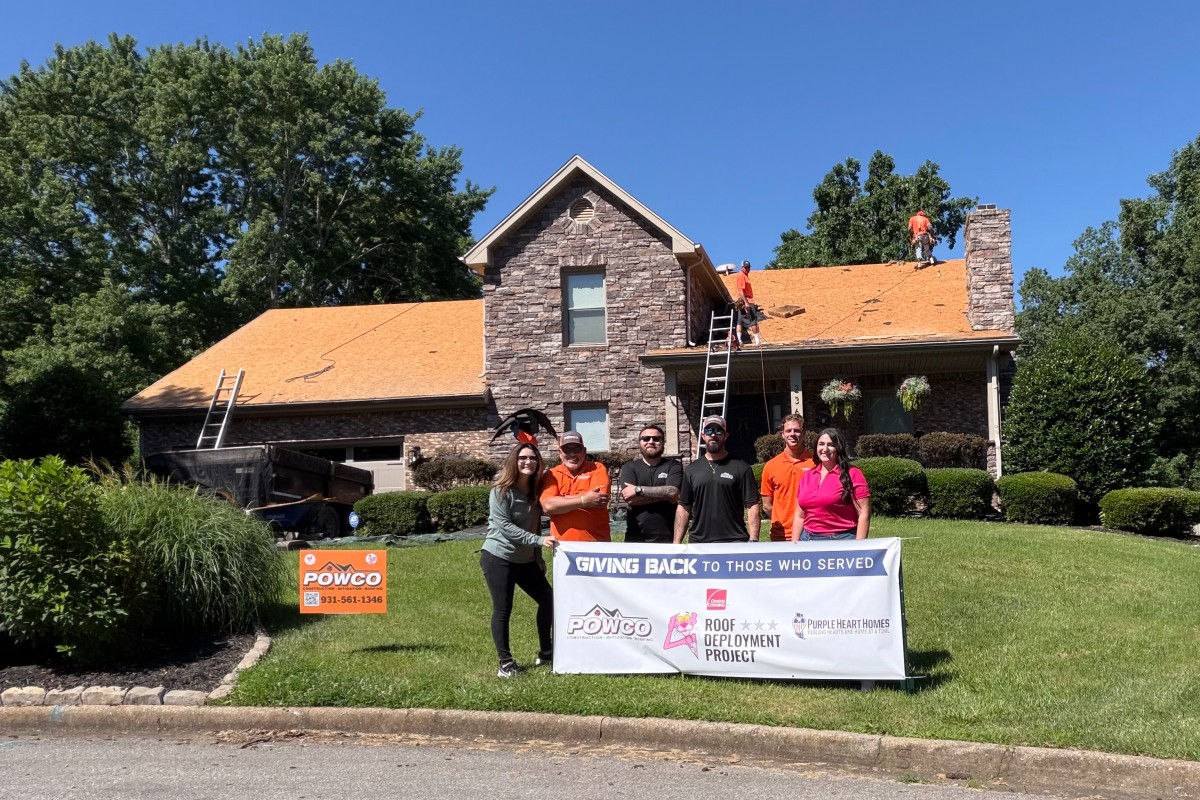 Volunteer group posing with a banner in front of a stone house while roof installers work on the orange roof on top of the house.