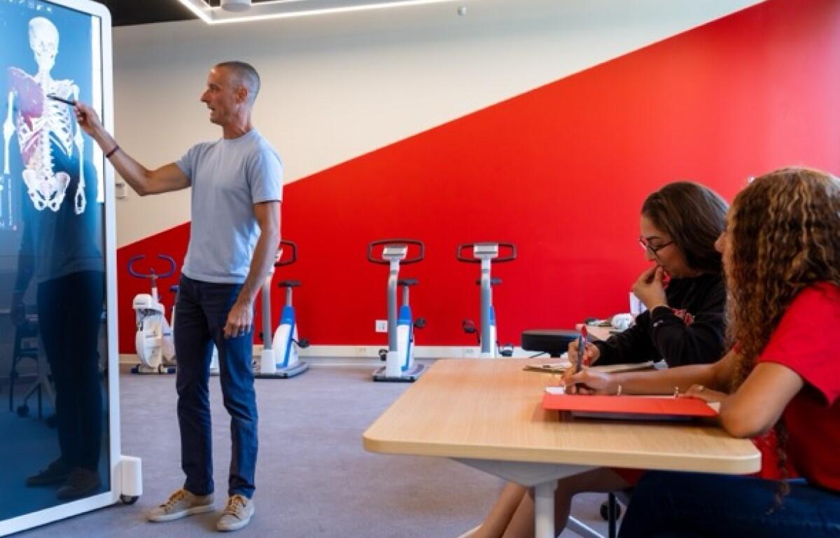 Instructor points to a full-body X-ray on a display mounted in a gym while explaining to two students taking notes at a nearby table with exercise bikes in the background.