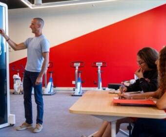Instructor points to a full-body X-ray on a display mounted in a gym while explaining to two students taking notes at a nearby table with exercise bikes in the background.