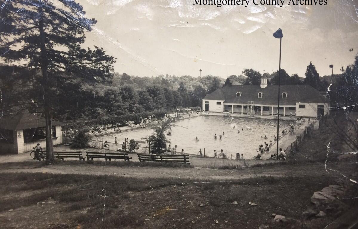 Black-and-white photo of a public swimming pool complex with a clubhouse, surrounded by trees and benches, and people swimming and relaxing around the pool.