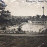 Black-and-white photo of a public swimming pool complex with a clubhouse, surrounded by trees and benches, and people swimming and relaxing around the pool.