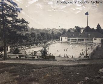 Black-and-white photo of a public swimming pool complex with a clubhouse, surrounded by trees and benches, and people swimming and relaxing around the pool.