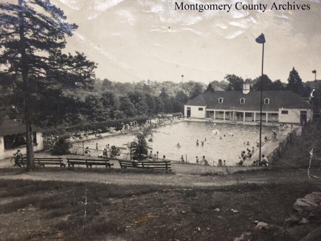 Black-and-white photo of a public swimming pool complex with a clubhouse, surrounded by trees and benches, and people swimming and relaxing around the pool.