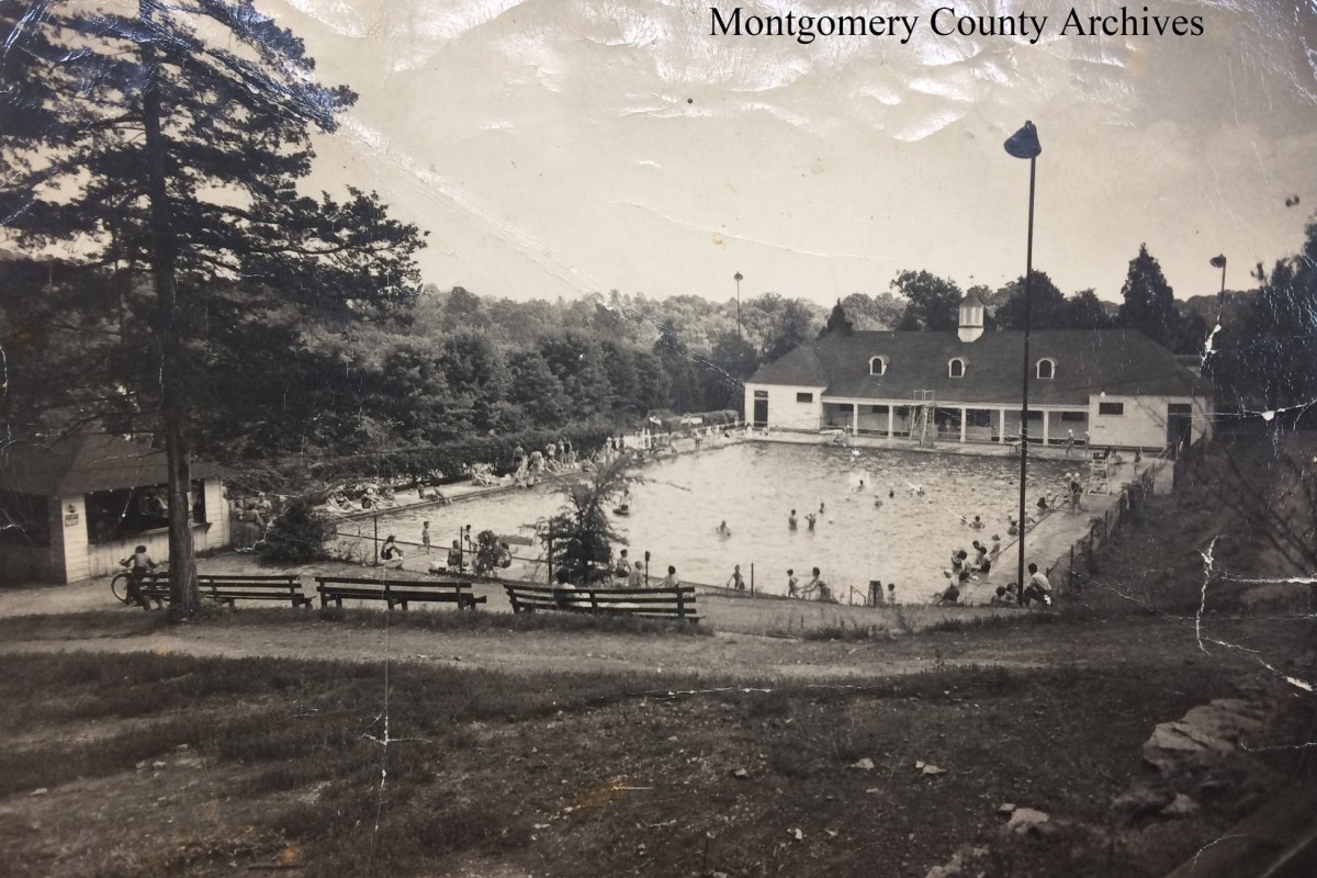 Black-and-white photo of a public swimming pool complex with a clubhouse, surrounded by trees and benches, and people swimming and relaxing around the pool.