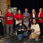 Eight people pose indoors with award plaques at a ceremony, smiling for the photo.