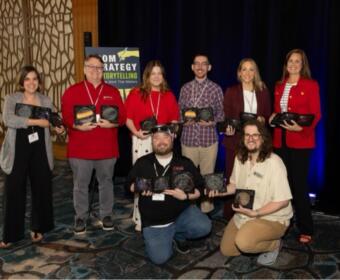 Eight people pose indoors with award plaques at a ceremony, smiling for the photo.