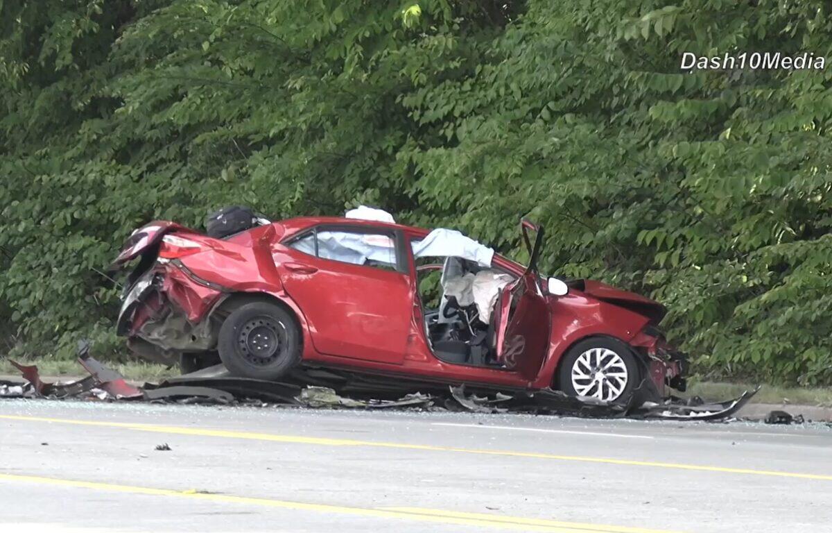 Red car severely damaged in a rollover/impact crash on a highway, with crushed front and rear ends and debris scattered nearby near a green treeline.