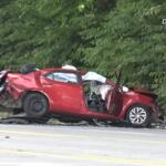 Red car severely damaged in a rollover/impact crash on a highway, with crushed front and rear ends and debris scattered nearby near a green treeline.