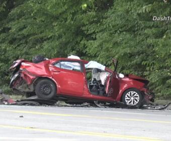 Red car severely damaged in a rollover/impact crash on a highway, with crushed front and rear ends and debris scattered nearby near a green treeline.