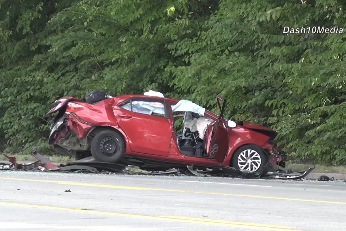 Red car severely damaged in a rollover/impact crash on a highway, with crushed front and rear ends and debris scattered nearby near a green treeline.