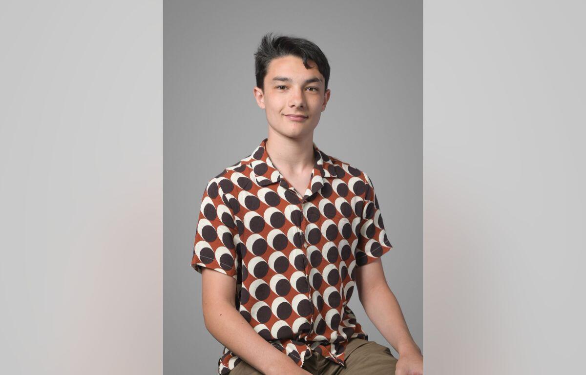 Young man sitting against a gray backdrop, wearing a brown and white circular-pattern shirt and khaki shorts, looking at the camera with a slight smile.