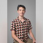 Young man sitting against a gray backdrop, wearing a brown and white circular-pattern shirt and khaki shorts, looking at the camera with a slight smile.