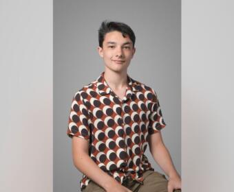 Young man sitting against a gray backdrop, wearing a brown and white circular-pattern shirt and khaki shorts, looking at the camera with a slight smile.