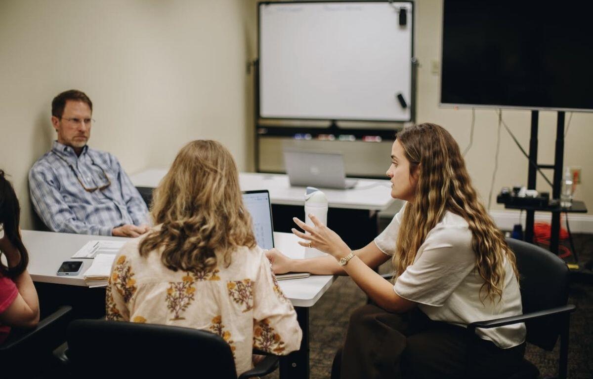 Woman with long hair gesturing while discussing with coworkers at a conference table in a meeting room, laptops open on the table.