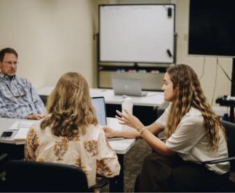 Woman with long hair gesturing while discussing with coworkers at a conference table in a meeting room, laptops open on the table.