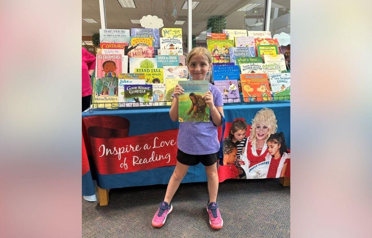 Young girl in a purple shirt and pink sneakers stands in front of a colorful children's book display, proudly holding up a book for the camera.