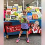 Young girl in a purple shirt and pink sneakers stands in front of a colorful children's book display, proudly holding up a book for the camera.