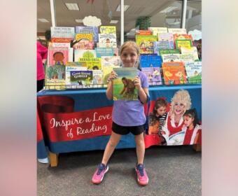 Young girl in a purple shirt and pink sneakers stands in front of a colorful children's book display, proudly holding up a book for the camera.