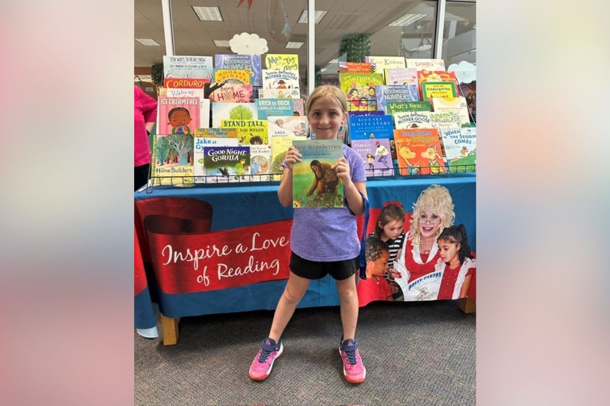 Young girl in a purple shirt and pink sneakers stands in front of a colorful children's book display, proudly holding up a book for the camera.