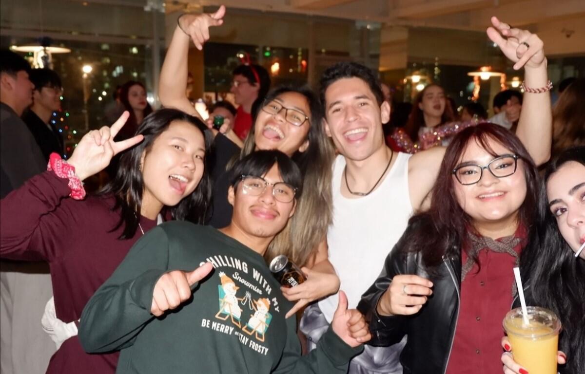 Group of friends at a party smiling and posing for the camera, some making peace signs, holding drinks and a microphone/struck pose.
