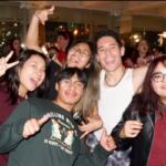 Group of friends at a party smiling and posing for the camera, some making peace signs, holding drinks and a microphone/struck pose.