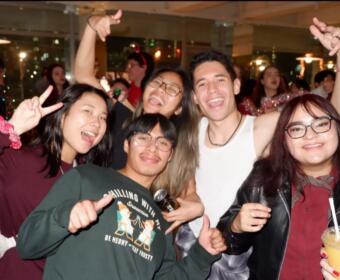 Group of friends at a party smiling and posing for the camera, some making peace signs, holding drinks and a microphone/struck pose.