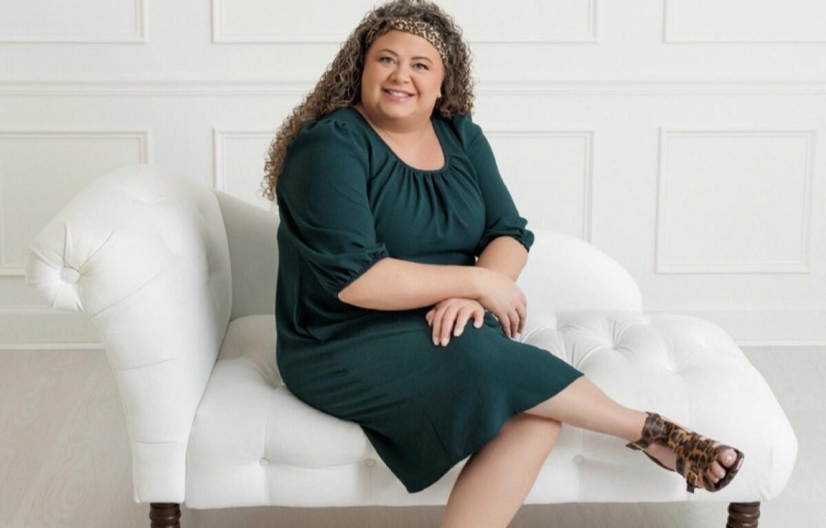 Smiling woman with curly hair in a dark green dress sits on a white tufted sofa, wearing leopard-print sandals.