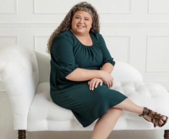 Smiling woman with curly hair in a dark green dress sits on a white tufted sofa, wearing leopard-print sandals.