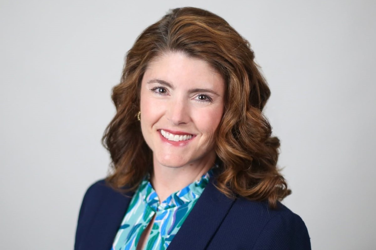 Professional headshot of a smiling woman with wavy brown hair, wearing a navy blazer and teal blouse against a light gray background.