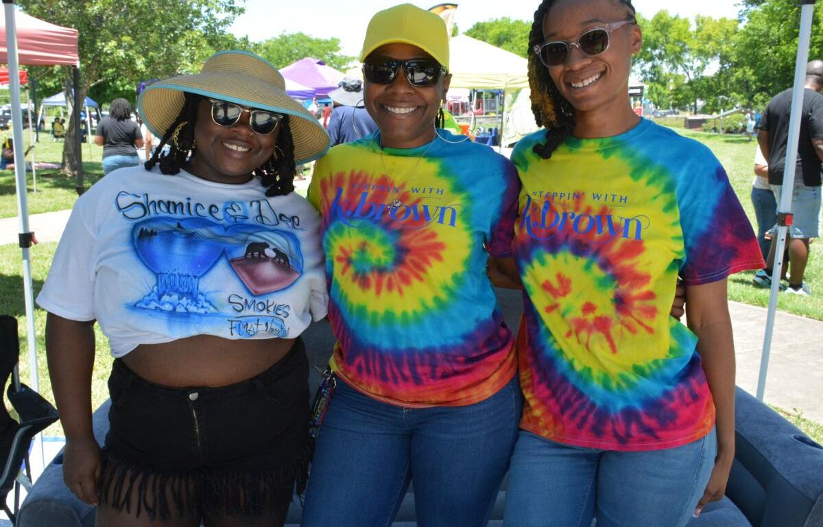 Three smiling women pose together at an outdoor festival under a canopy; two wear tie-dye shirts and sunglasses, one wears a straw hat with a graphic tee and fringe shorts.