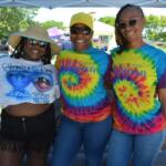 Three smiling women pose together at an outdoor festival under a canopy; two wear tie-dye shirts and sunglasses, one wears a straw hat with a graphic tee and fringe shorts.