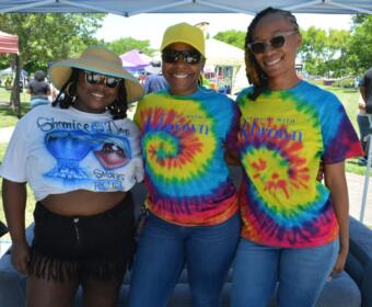 Three smiling women pose together at an outdoor festival under a canopy; two wear tie-dye shirts and sunglasses, one wears a straw hat with a graphic tee and fringe shorts.