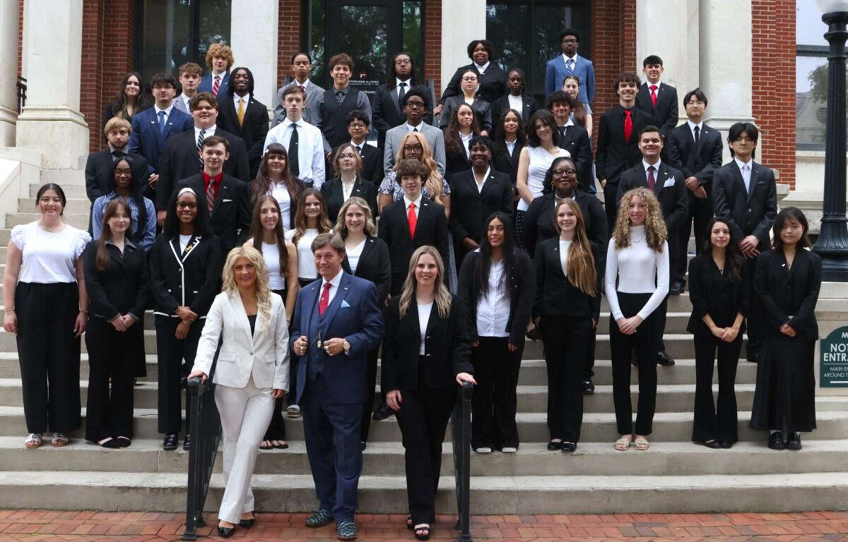 Group of diverse students in formal attire posing on building steps with two adults at the front center in suits and dresses.
