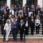 Group of diverse students in formal attire posing on building steps with two adults at the front center in suits and dresses.