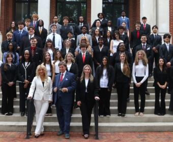 Group of diverse students in formal attire posing on building steps with two adults at the front center in suits and dresses.